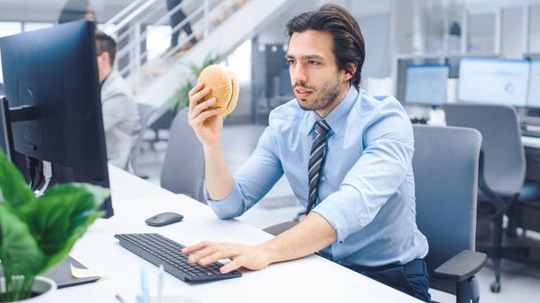 In the Bright Modern Office Young Businessman Eats Hamburger Sitting at His Desktop Computer, Prepared to Bite it and Continue to Work During His Lunch. In the Background Colleagues Working - Foto, immagini