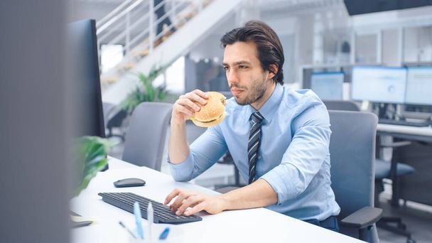 In the Bright Modern Office Young Businessman Eats Hamburger Sitting at His Desktop Computer, He Chewing the Bun and Continues to Work During His Lunch. In the Background Colleagues Working - Foto, Bild