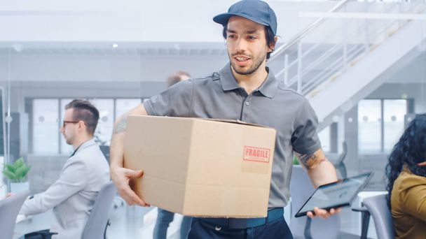 Happy Delivery Man Wals in Corporate Office with Tablet Computer and Delivers Cardboard Package to a Worker. Big Bright Modern Business Company Office with Professional Businesspeople Working - Фото, зображення