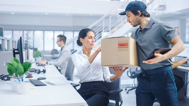Happy Delivery Man Enters Corporate Office, Hands Package to a Beautiful Businesswoman. He Holds Tablet Computer. Big Bright Modern Business Company Office with Professional Businesspeople Working - 写真・画像