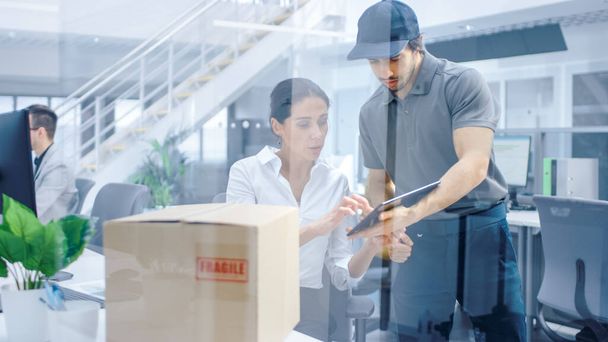 Happy Delivery Man Enters Corporate Office, Hands Package to a Beautiful Businesswoman, She signs Her Signature on the Tablet Computer. Big Bright Modern Business Company Office with Professionals. - 写真・画像