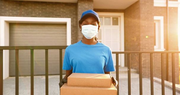 Portrait of African American young woman courier in medical mask, gloves and hat standing at house in street and holding carton parcels. Female delivery worker in uniform with boxes. Quarantine time. - Photo, image