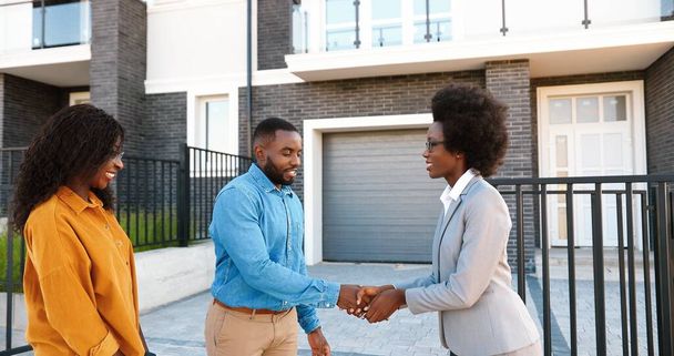Female real-estate selling house to African American happy couple and passing keys. Outdoors. Cheerful just-married man and woman smiling and buying home at outskirt. Getting dwelling in suburb. - Foto, Bild