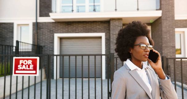 African American young stylish female real-estate agent in glasses at outskirt neighborhood and talking on mobile phone. Businesswoman looking for house and speaking on cellphone. Dwelling for sale. - Фото, изображение