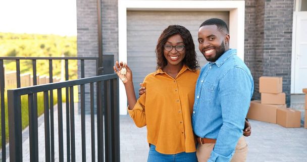 Portrait of happy young African American man and woman smiling to camera and demonstrating key of new home. Outside. Buying real-estate. Moving in concept. Blurred. Cheerful couple settling in. - Fotografie, Obrázek