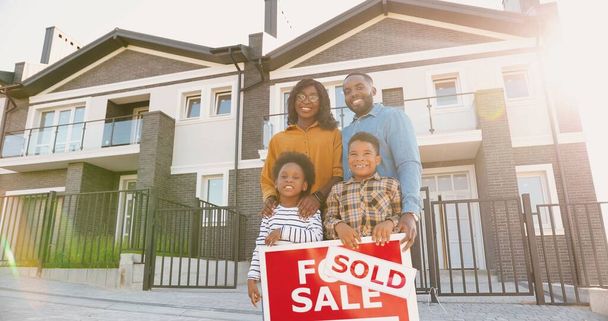 Portrait of happy African American family with small children standing at new house at suburb and smiling with sold table in hands. Cheerful parents and kids moving in new home. Settlement in outskirt - Foto, afbeelding
