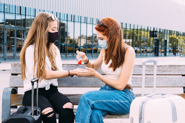 две молодые туристки, сидящие на скамейке возле вокзала, надевают антибактериальный гель - Фото, изображение