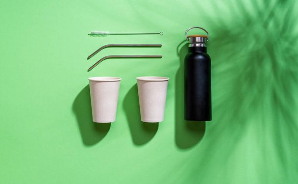 Вид сверху с непластиковыми чашками, металлическими соломинками и термосом. Устойчивые питьевые контейнеры изолированы на зеленом фоне. Бумажные стаканчики и термос для устойчивых путешествий. - Фото, изображение