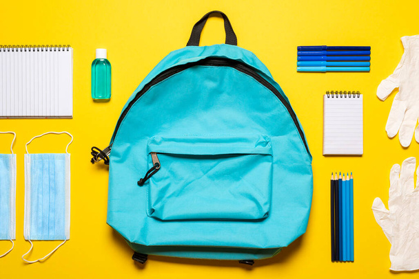 Back to school flat lay with face masks, gloves,  hand sanitizer, and school supplies. Blue backpack, hygiene objects, and school supplies isolated on a yellow background. - Photo, Image