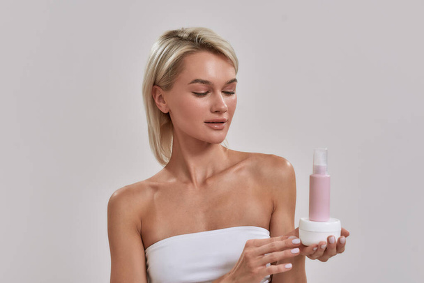 Portrait of young woman with perfect glowing skin holding jar and bottle of different cosmetic skincare products and looking at it while posing isolated over grey background - Photo, image