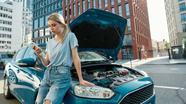 Attractive young woman holding phone, calling emergency car service while standing, leaning on her broken car with open hood on the city street - Photo, Image