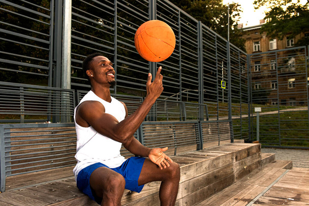 Jonge Afro-Amerikaanse basketbalspeler in wit t-shirt en blauwe shorts zittend op basketbalveld, draaiende basketbal op vingers, bedacht, sport concept. - Foto, afbeelding
