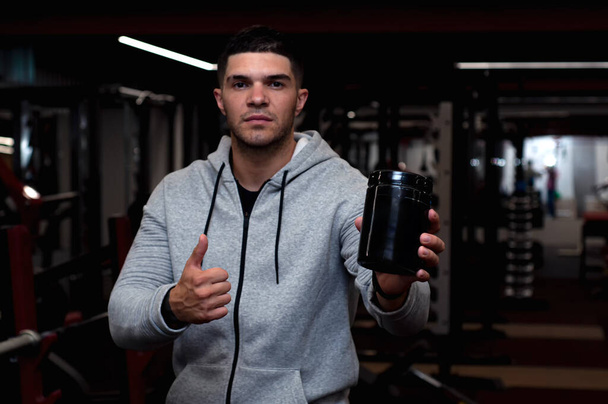 Young athletic guy holding a jar of sports nutrition, on gym background - Фото, зображення