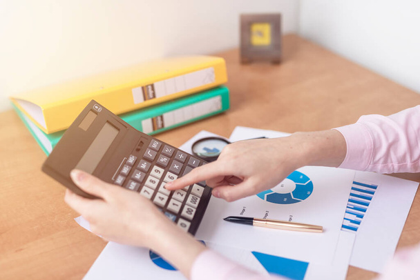 female accountant or banker making calculations sitting at office table with documents with tables on desk. Savings, finances and economy concept, counting on calculator - Foto, Imagem