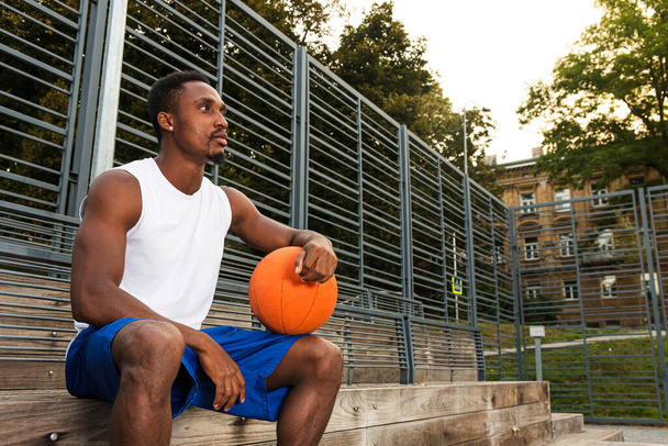 Joven jugador de baloncesto afroamericano en camiseta blanca y pantalones cortos azules en la cancha de baloncesto sentado, sosteniendo el baloncesto, concebido, concepto deportivo - Foto, imagen