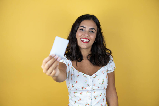Mooie vrouw over gele achtergrond glimlachen en het houden van witte kaart - Foto, afbeelding