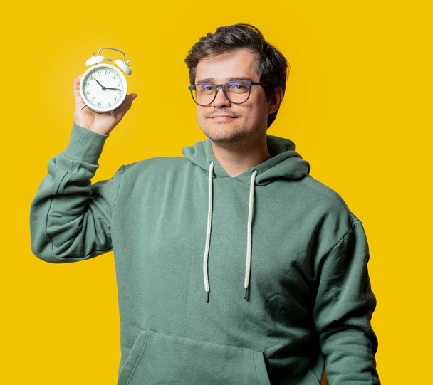 man in groene hoodie houden wekker op gele achtergrond - Foto, afbeelding