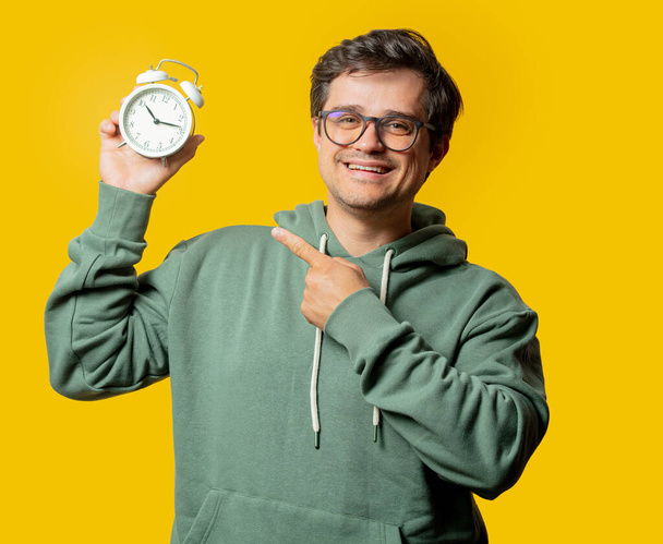 hombre con capucha verde mantenga el reloj despertador sobre fondo amarillo - Foto, Imagen