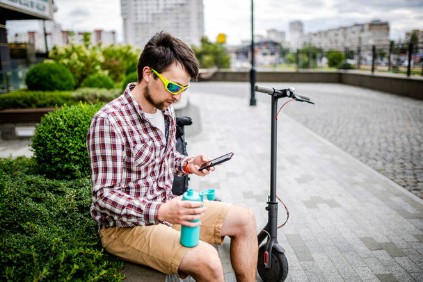 白人男性は、電動スクーターの近くの市内のベンチに座っている間、スマートフォン上の現代的な技術インターネットと携帯電話通信を使用します。生態系と環境に優しい都市交通. - 写真・画像