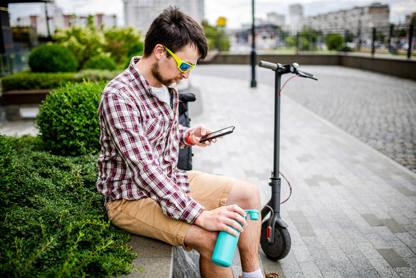 Modern ecological clean transport theme. A man resting on a bench and holding a smart phone in his hand after riding an eco electric kick scooter in the city. Male riding a scooter checking messages. - 写真・画像
