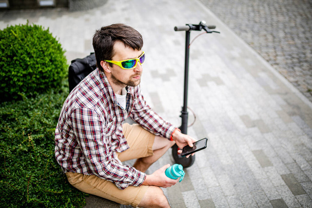 Modern ecological clean transport theme. A man resting on a bench and holding a smart phone in his hand after riding an eco electric kick scooter in the city. Male riding a scooter checking messages. - Zdjęcie, obraz