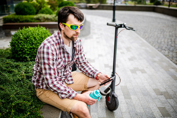 Mann mit E-Roller entspannt sich und nutzt Smartphone auf Bank in der Stadt. Moderner Mann mit Elektroroller sitzt auf Bank im Park, während er mit seinem Handy chattet. Student mit Ökotransport hält Handy in der Hand. - Foto, Bild