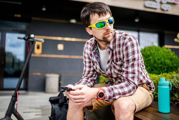 A kaukázusi férfi modern technológiákat használ Internet és mobilkommunikáció okostelefonon, miközben a padon ül a városban, közel az elektromos robogóhoz. Ökológia és környezetbarát városi közlekedés. - Fotó, kép