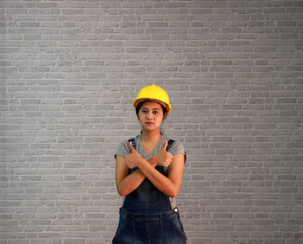 Techniker Frau ware gelber Helm mit grauem T-Shirt und Jeans Schürze Kleid stehend und Daumen nach oben zwei Hände auf grauem Backsteinmuster Hintergrund. - Foto, Bild
