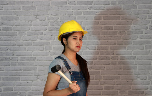 Технік жінка поставляє жовтий шолом з сірою футболкою та джинсовою сукнею з фартухами, що стоїть та гумовим молотком у руці на сірому фоні візерунка з цегли
. - Фото, зображення