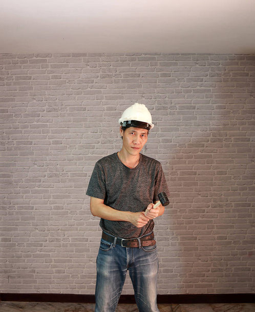 Technicus man ware witte helm met donkergrijze T-shirt en spijkerbroek staande met rubberen hamer in twee handen op grijze baksteen patroon achtergrond. - Foto, afbeelding