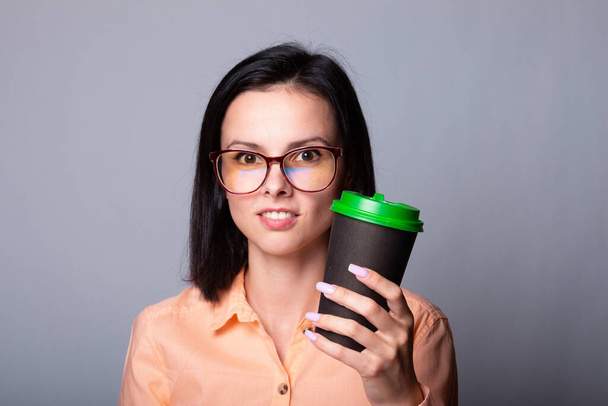 dívka student v oranžové košili a brýle drží šálek kávy v ruce, šedé pozadí - Fotografie, Obrázek