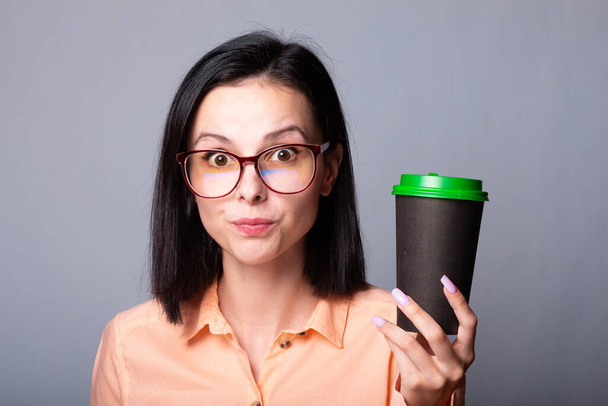 woman in an orange shirt and glasses holding a cup of coffee in her hands, gray background - 写真・画像