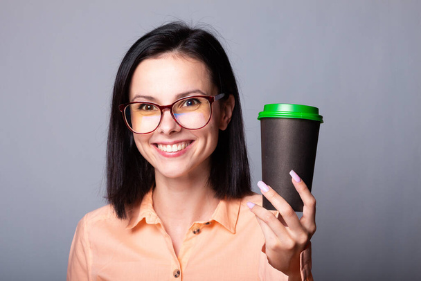 woman in an orange shirt and glasses holding a cup of coffee in her hands, gray background - Photo, image