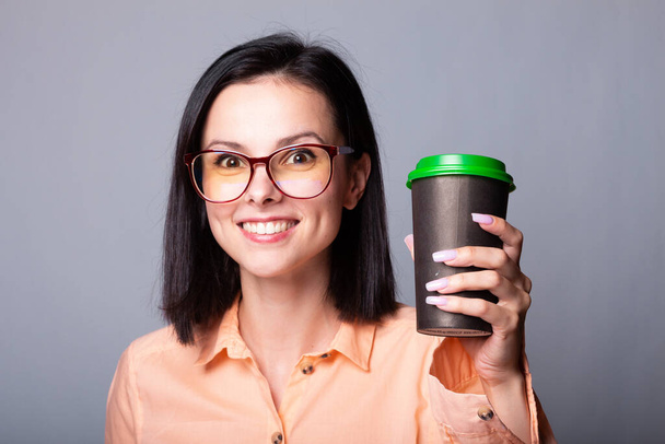woman in an orange shirt and glasses holding a cup of coffee in her hands, gray background - Foto, immagini