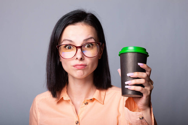 woman in an orange shirt and glasses holding a cup of coffee in her hands, gray background - Foto, imagen