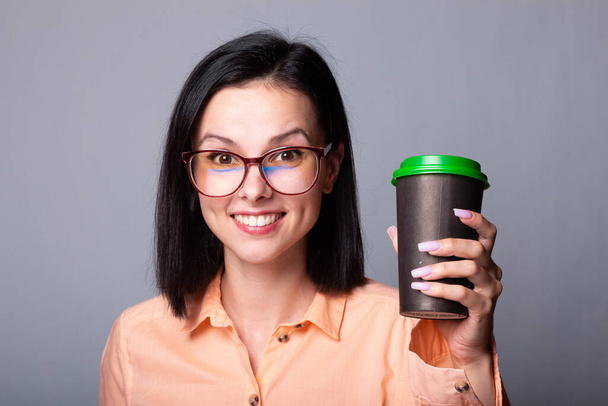 woman in an orange shirt and glasses holding a cup of coffee in her hands, gray background - Foto, Bild