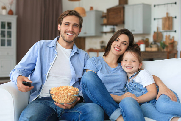 Happy family with child sitting on sofa watching tv and eating popcorn, young parents embracing daughter relaxing on couch together - Фото, изображение