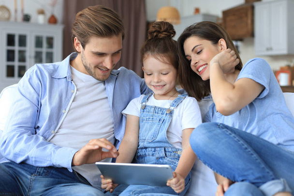 Positive friendly young parents with smiling little daughter sitting on sofa together answering video call on digital tablet while relaxing at home on weekend - Fotoğraf, Görsel