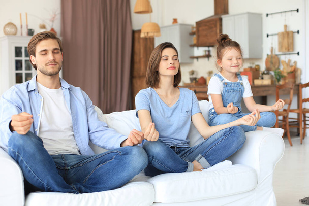 Calm young family with little daughter sit on couch practice yoga together, happy parents with small preschooler girl child rest on sofa meditate relieve negative emotions on weekend at home - Foto, Imagem