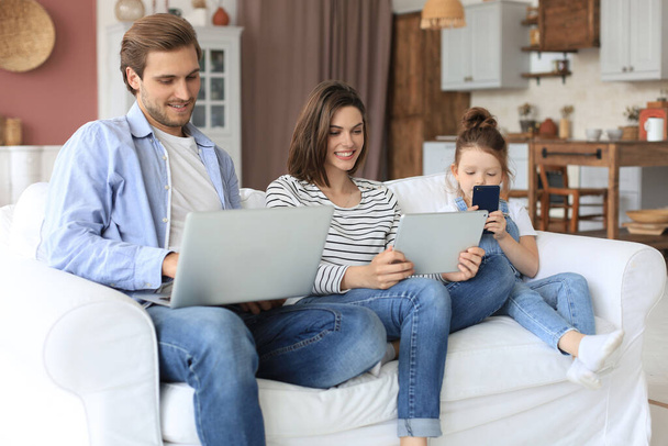 Father, mother and daughter using electronic devices sitting on sofa at living room - Foto, afbeelding