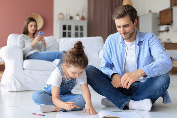 Happy father smilling daughter lying on warm floor enjoying creative activity, drawing pencils coloring pictures in albums, mother resting on couch, family spend free time together - Photo, Image