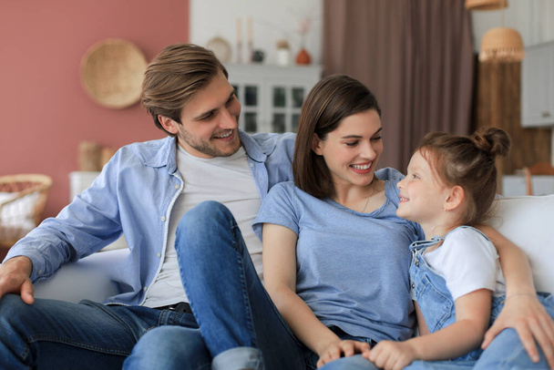 Positive friendly young parents with smiling little daughter sitting on sofa together while relaxing at home on weekend - Photo, image
