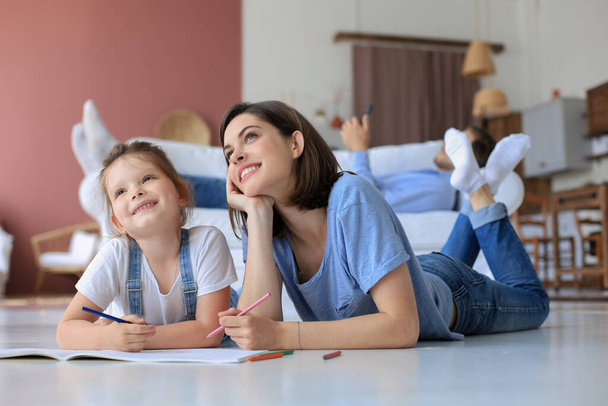 Happy mother smilling daughter lying on warm floor enjoying creative activity, drawing pencils coloring pictures in albums, father resting on couch, family spend free time together - Foto, Bild