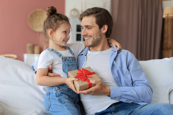 Cute kid daughter make surprise to daddy, little girl presents gift box to father sit on sofa. Father's day - Фото, изображение