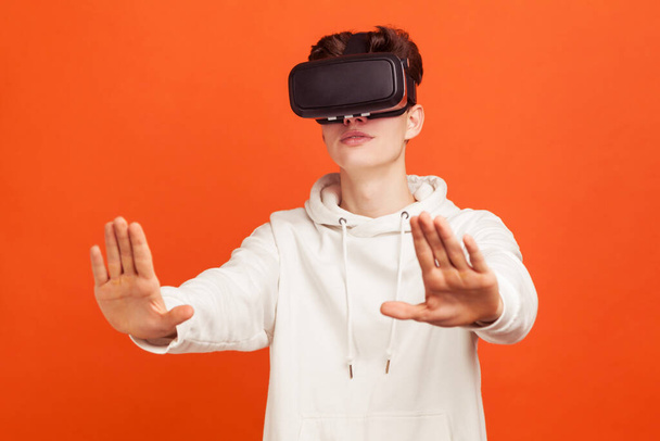 Excited young man in casual sweatshirt in virtual reality headset conducting online conference, colleagues reviewing and discussing 3d project. Indoor studio shot isolated on orange background - Foto, Imagem