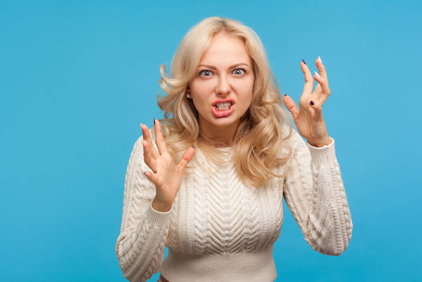 Aggressive stressed blond woman arguing and becoming mad clenching teeth, crazy furious lady in depression. Indoor studio shot isolated on blue background - Фото, изображение