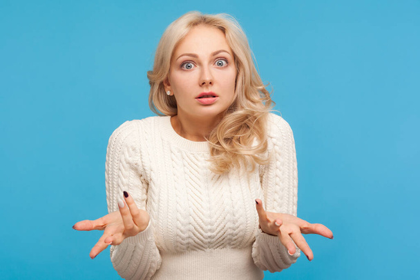 Unhappy frustrated woman with curly blond hair spreading hands with shock and misunderstanding. How could you? Indoor studio shot isolated on blue background - 写真・画像