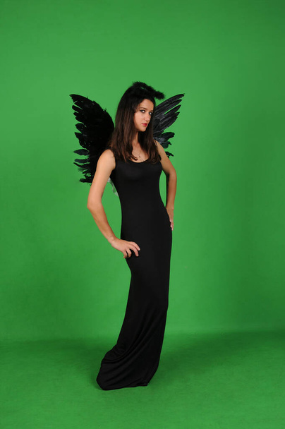 Hermosa joven morena en traje de ángel negro con alas sobre fondo verde. Traje de fiesta de Halloween - Foto, Imagen