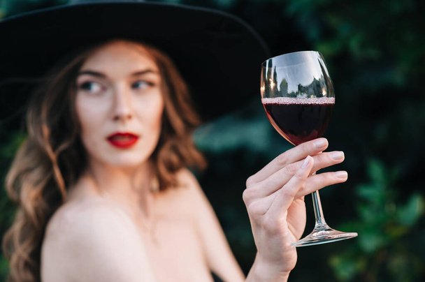 Ukrainian girl in a black dress outdoors in a hat with a glass of red wine - Photo, image