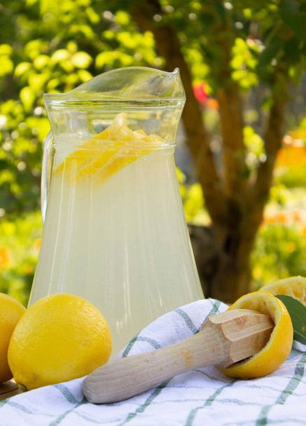 Jarra de vidrio / jarra con limonada en una mesa de madera en el jardín. Limones maduros, pistilo cítrico sobre toalla blanca - Foto, Imagen